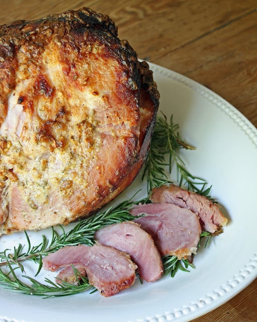 Rosemary and mustard crusted baked ham on a white platter garnished with branches of fresh rosemary and showing a few slices.