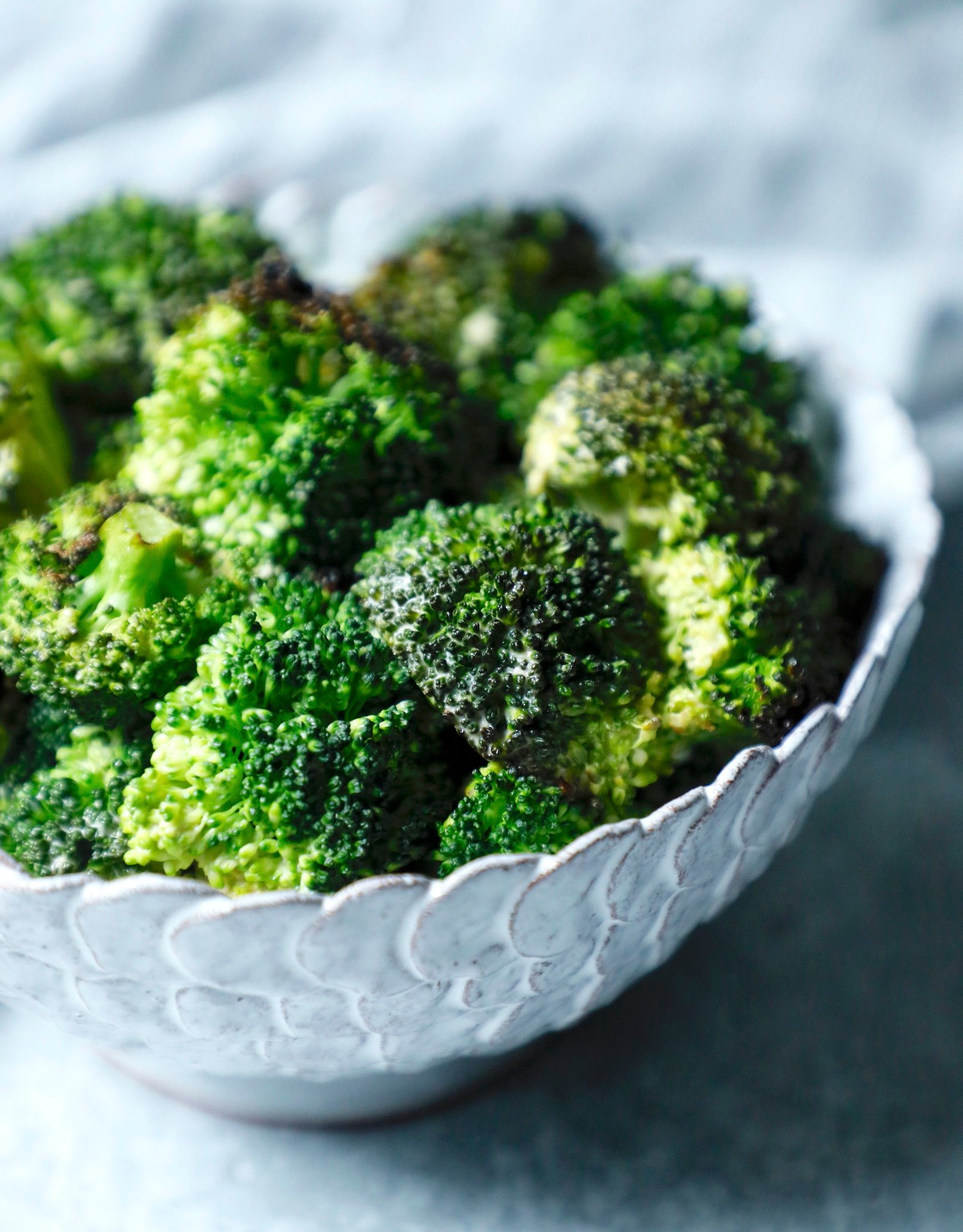 Front view of a white pottery bowl on a grey background filled with florets of sriracha roasted broccoli.