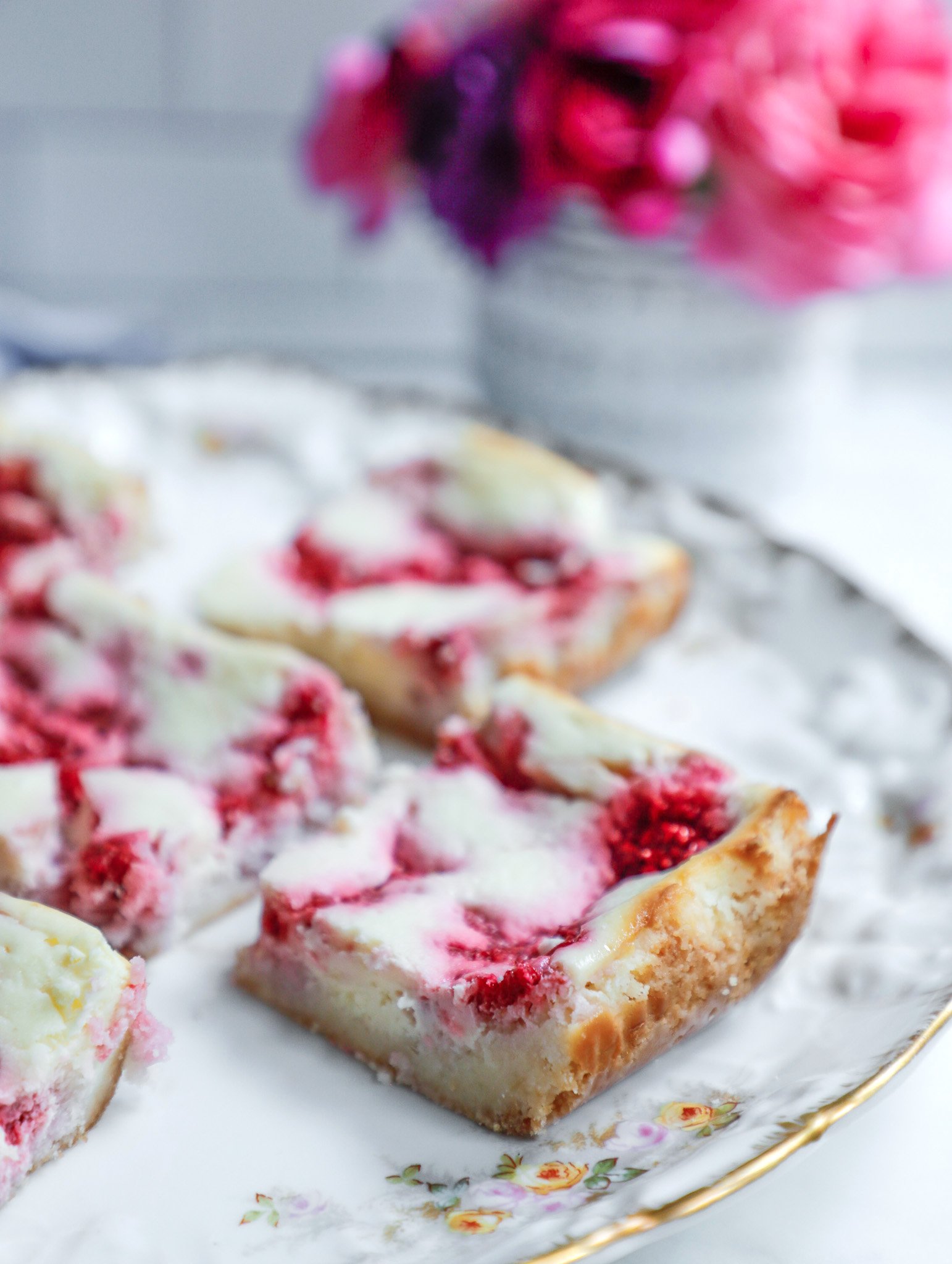 Raspberry cheesecake bars on a vintage platter with a white vase full of pink flowers in the background