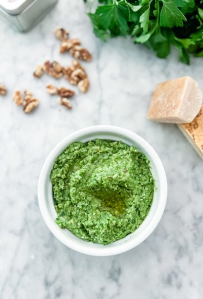 garlic scape pesto in a white bowl on a marble table. Surrounded by parmesan, walnuts and parsley arranged around the bowl