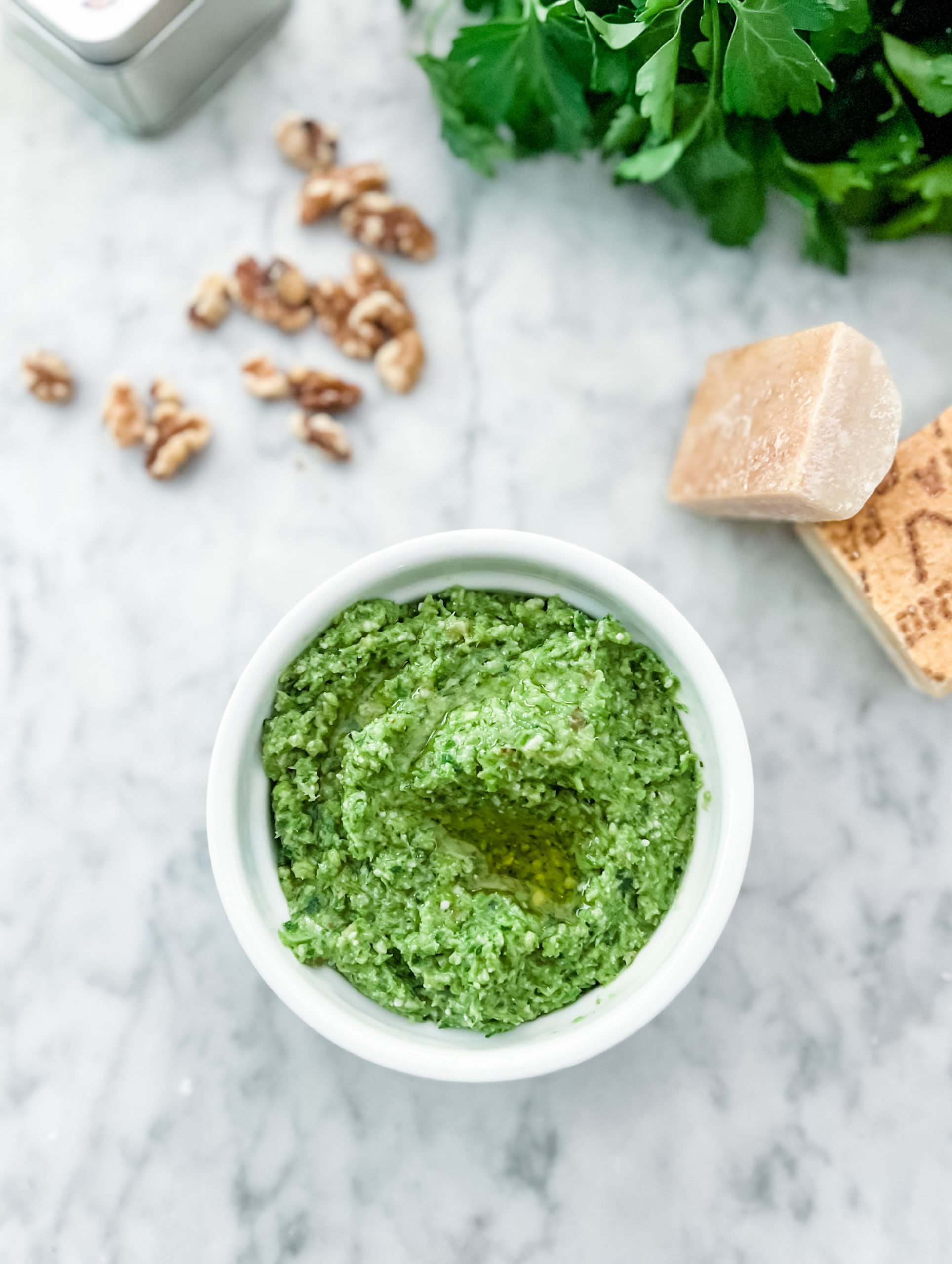 garlic scape pesto in a white bowl on a marble table. Surrounded by parmesan, walnuts and parsley arranged around the bowl