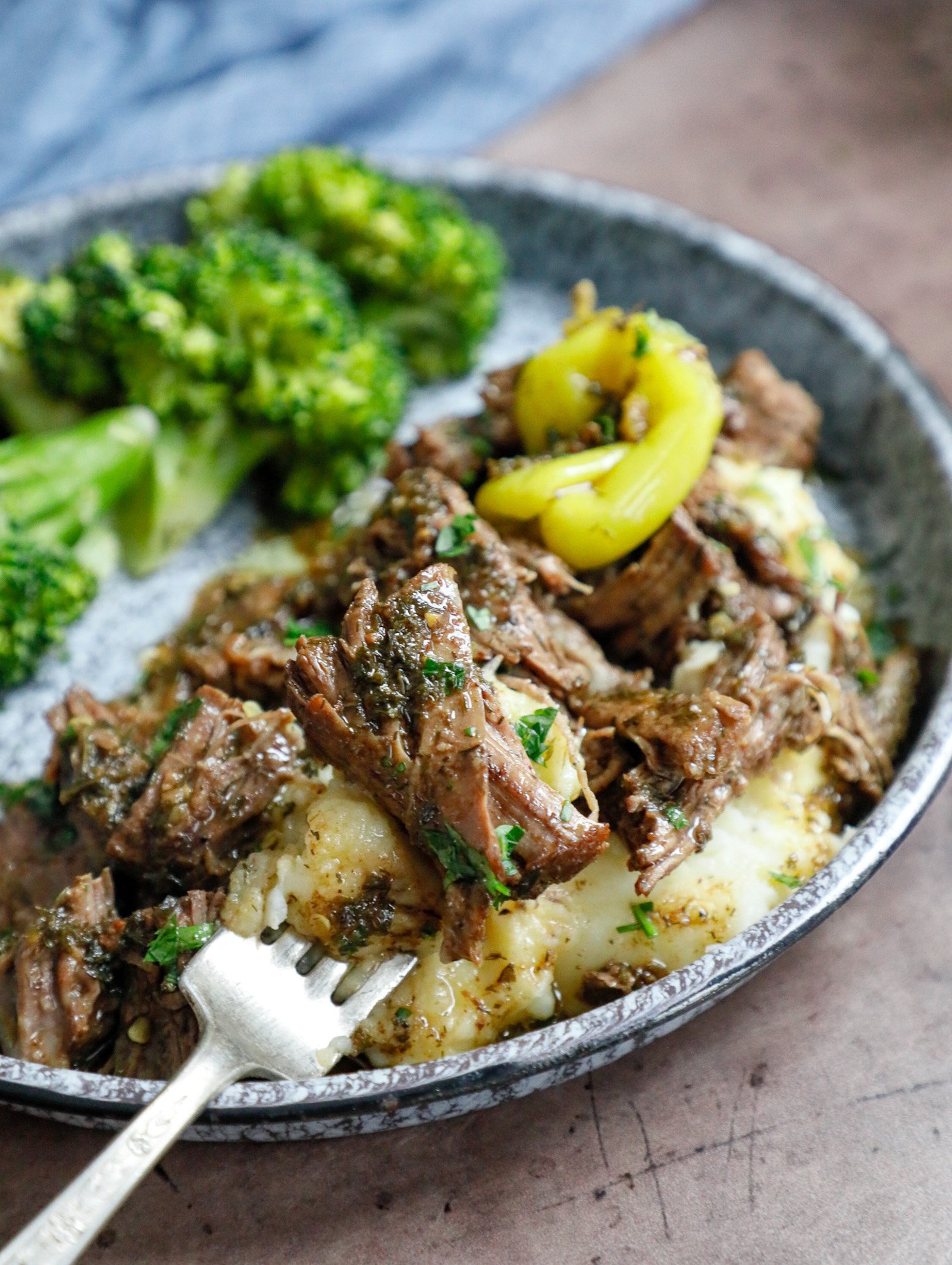 A forkful of shredded Keto Mississippi Pot Roast served over cauliflower mash on a blue enamelware plate with broccoli on the side