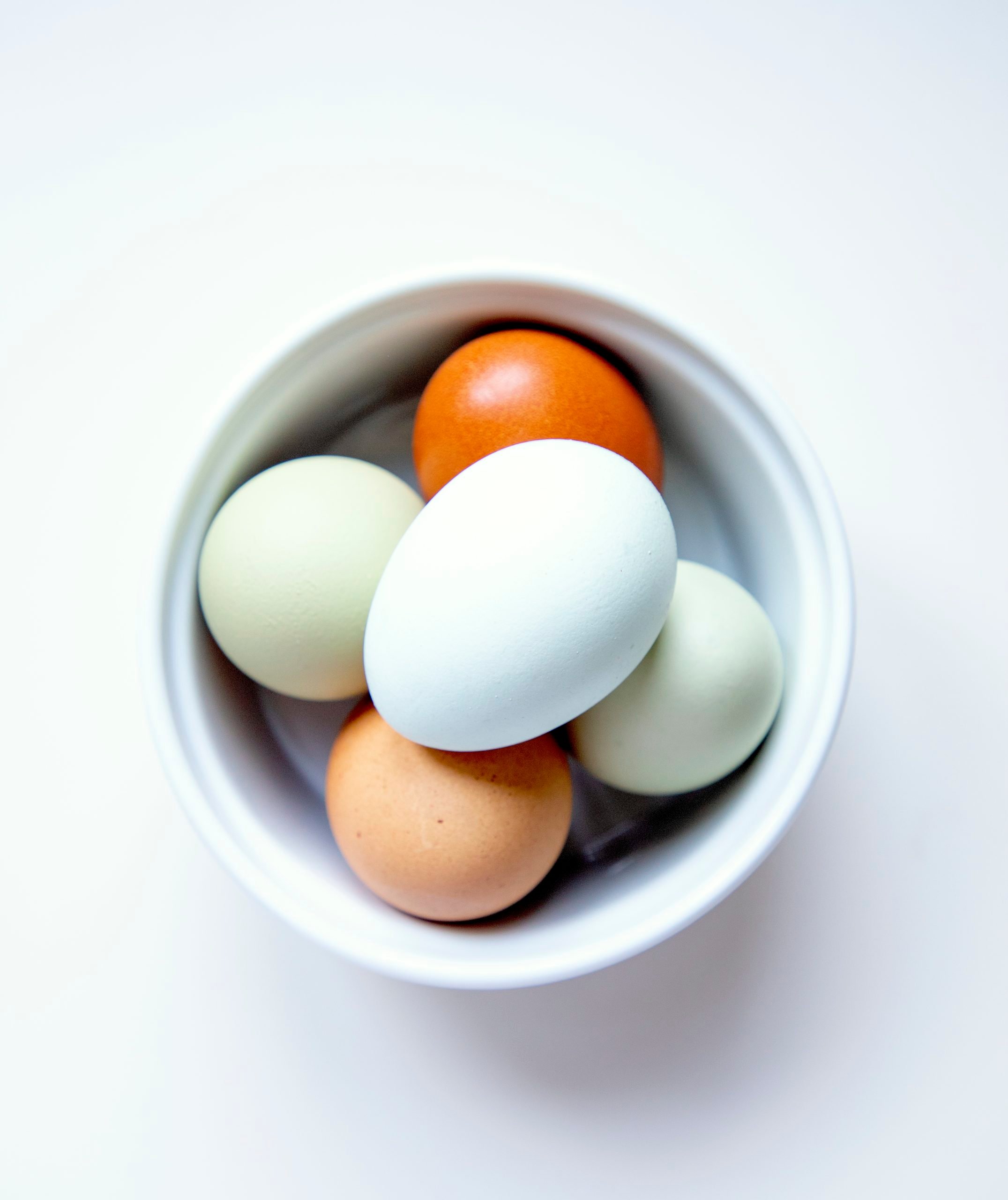 A white bowl with 5 whole eggs in it, the shells are all different colors - brown, light brown, white, and two shades of green